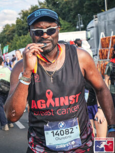 Timi wearing a black hat and a black ABC running vest, biting his Berlin Marathon 2025 medal