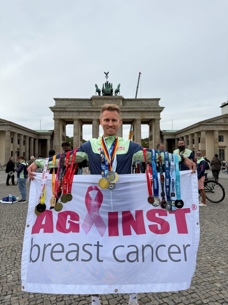 Jamie holding an Against Breast Cancer in front of the Brandenburg Gate after the Berlin Marathon with all of the medals he has for his races in 2025.