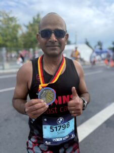 Srini wearing a black ABC running jersey and sunglasses, holding his medal and giving a thumbs up to the camera.
