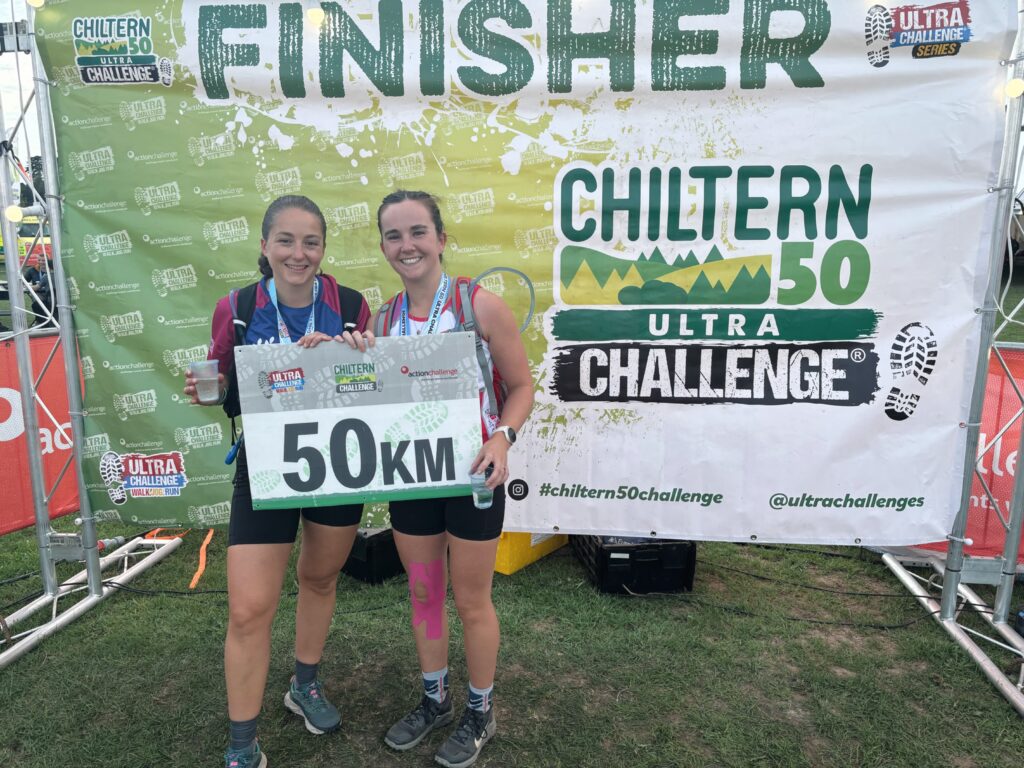 Helen and Polly holding a '50 km' sign in front of a finishers' banner after completing Chiltern 50 Challenge