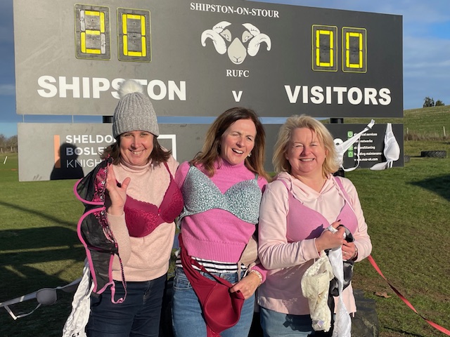 Three smiling women from Shipston-on-Stour Rugby Club stand arm-in-arm on a rugby pitch, wearing pink bras over their clothes as part of a light-hearted fundraiser. A scoreboard behind them reads ‘Shipston v Visitors 20–0’ on a bright, sunny day.