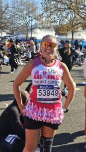 Lisa wearing a bright pink Against Breast Cancer running vests in a post‑race area surrounded by other participants. The outfit includes a skirt decorated with many small pink ribbons.