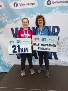 Emma and Mandy at the finish line, holding up cardboard banners saying "21 KM" and "Half Marathon Finisher" respectively.