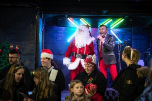 Brian dressed as Santa on stage with a man in a silver jacket and red trousers.