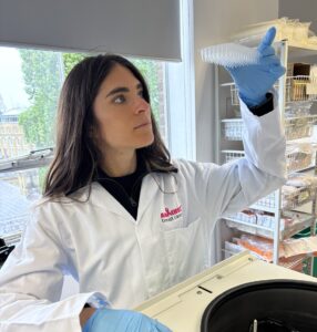 Ludovica in a lab coat and gloves holds up a multi-well plate to the light in a laboratory, with racks of scientific supplies and equipment visible behind.