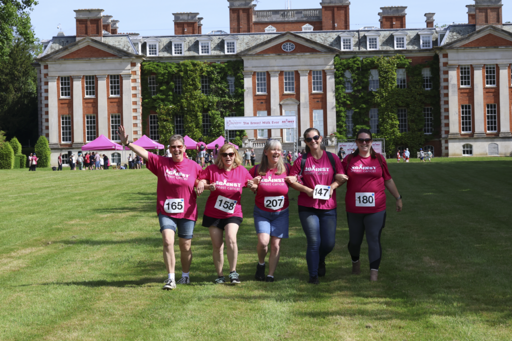 Five participants in pink Against Breast Cancer t-shirts walk across a lawn towards the camera in front of Hursley House, a large historic building, with event tents and a banner in the background.