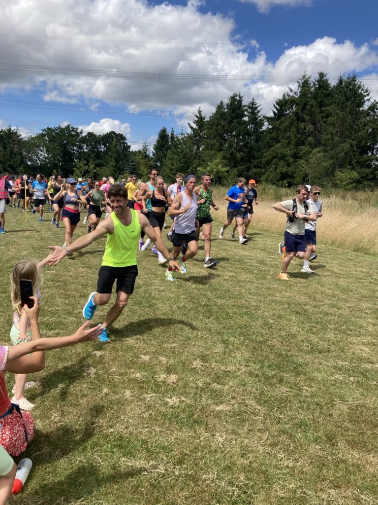 A group of runners running through a field at the Well Wellow 10k. In the foreground a man in a florescent running vest high fives a small girl.