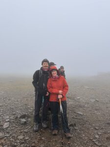 Pam and another male participant in waterproof jackets stand with trekking poles on a rocky summit shrouded in fog during the Yorkshire Three Peaks challenge.