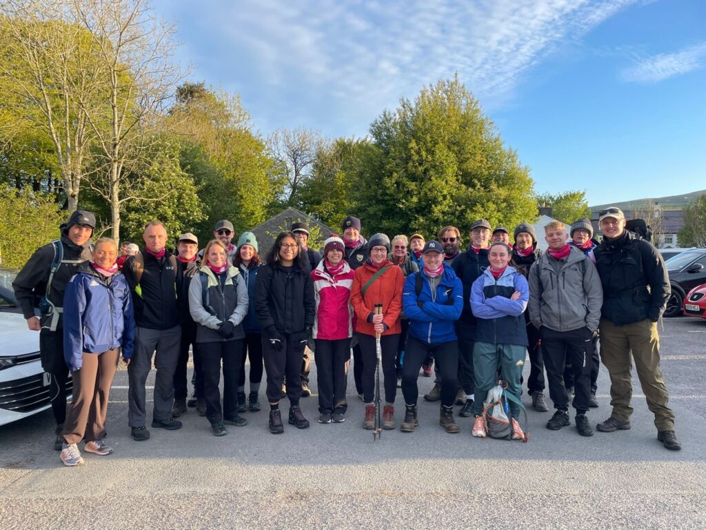 Pam with 22 of her friends and family in outdoor walking gear pose for a group photo at the start of the Yorkshire Three Peaks fundraising challenge for Against Breast Cancer.
