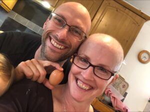 Rebecca and her husband, holding an egg which appears to symbolise her balding head from breast cancer treatment.